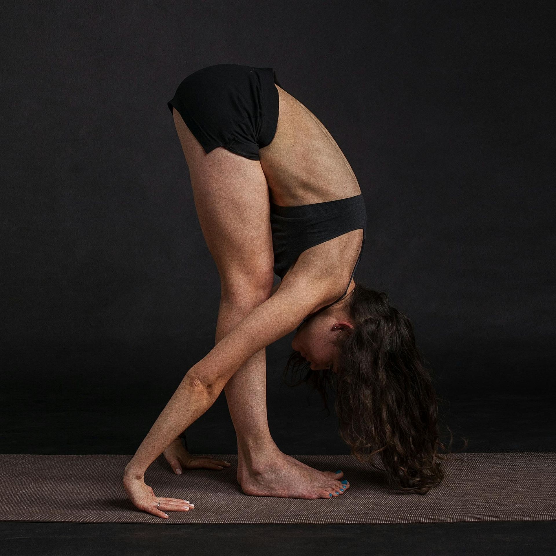 Woman in yoga pose, head down, hands on ground, legs crossed, black outfit, on mat.