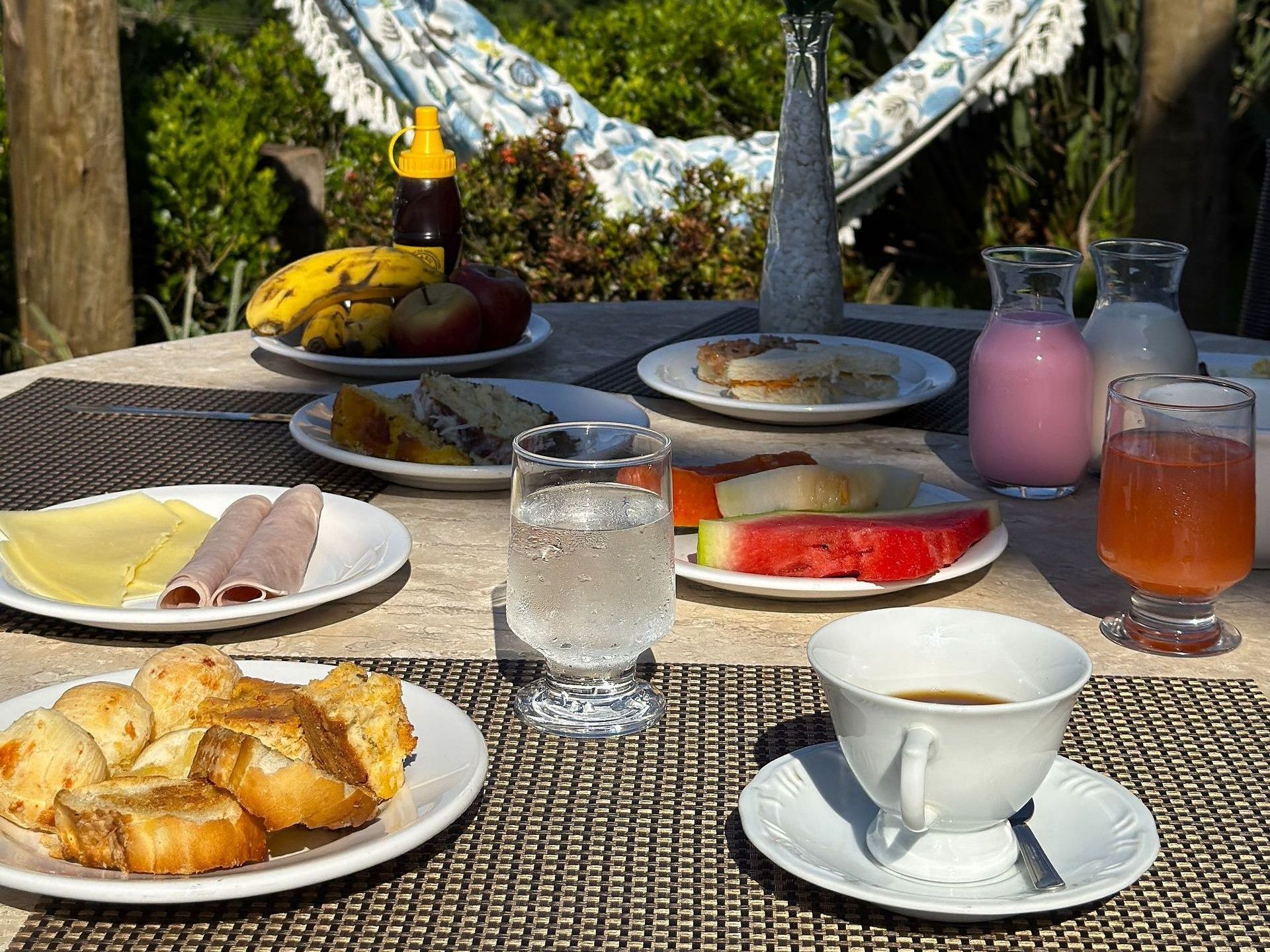 Uma mesa coberta com pratos de comida e bebidas com uma rede ao fundo. Hotel Quadrado