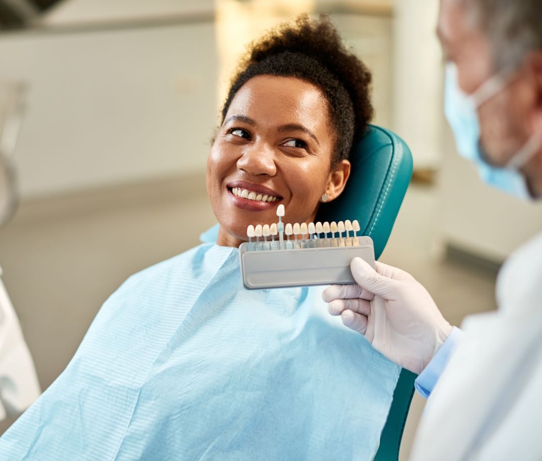 A woman is sitting in a dental chair while a dentist holds a tooth color chart.