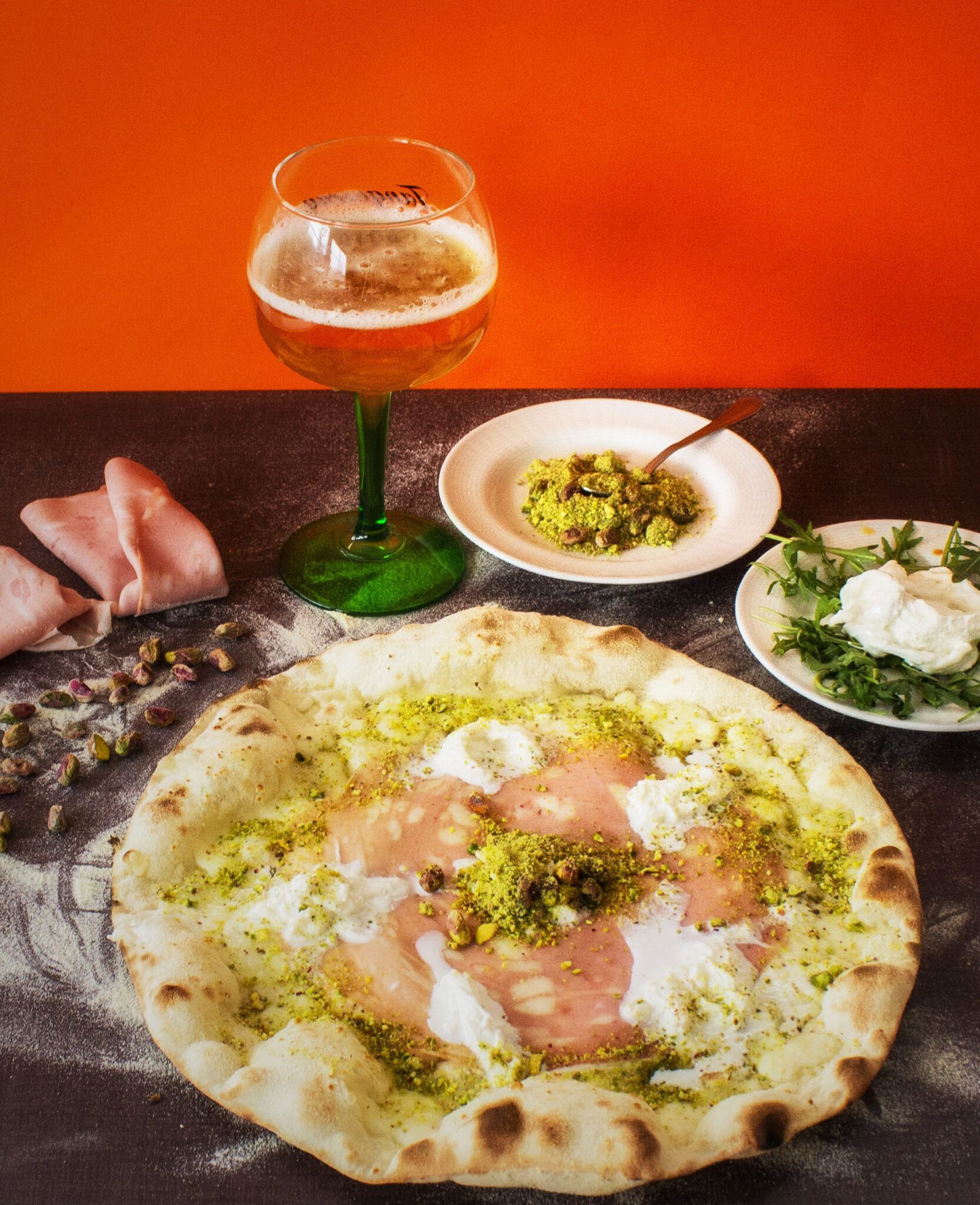 Pizza e antipasti su un tavolo scuro con una bevanda dal gambo verde contro una parete arancione.