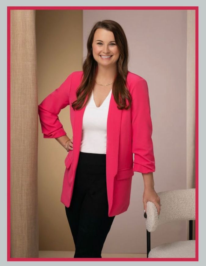 A woman wearing a pink jacket and black pants is standing next to a chair.