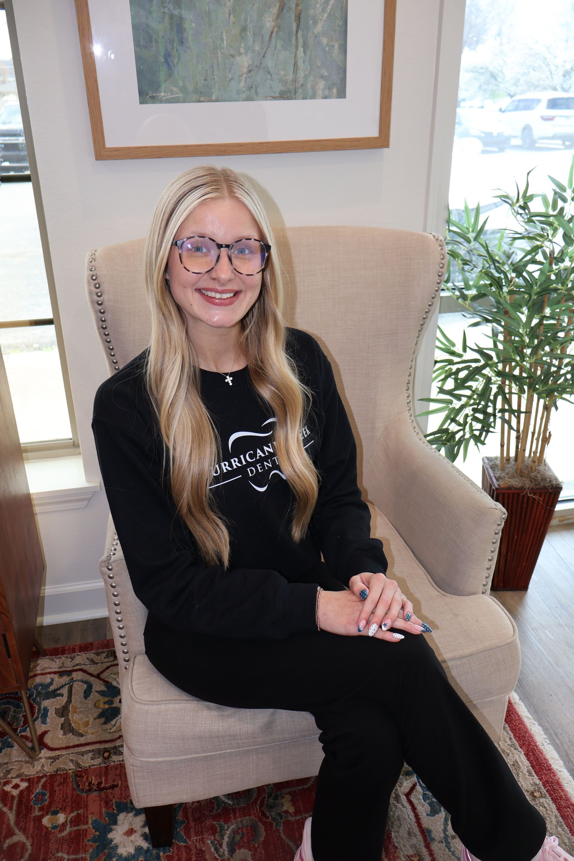 A person with long blonde hair and glasses sits in a beige armchair, smiling while wearing a black long-sleeved shirt.