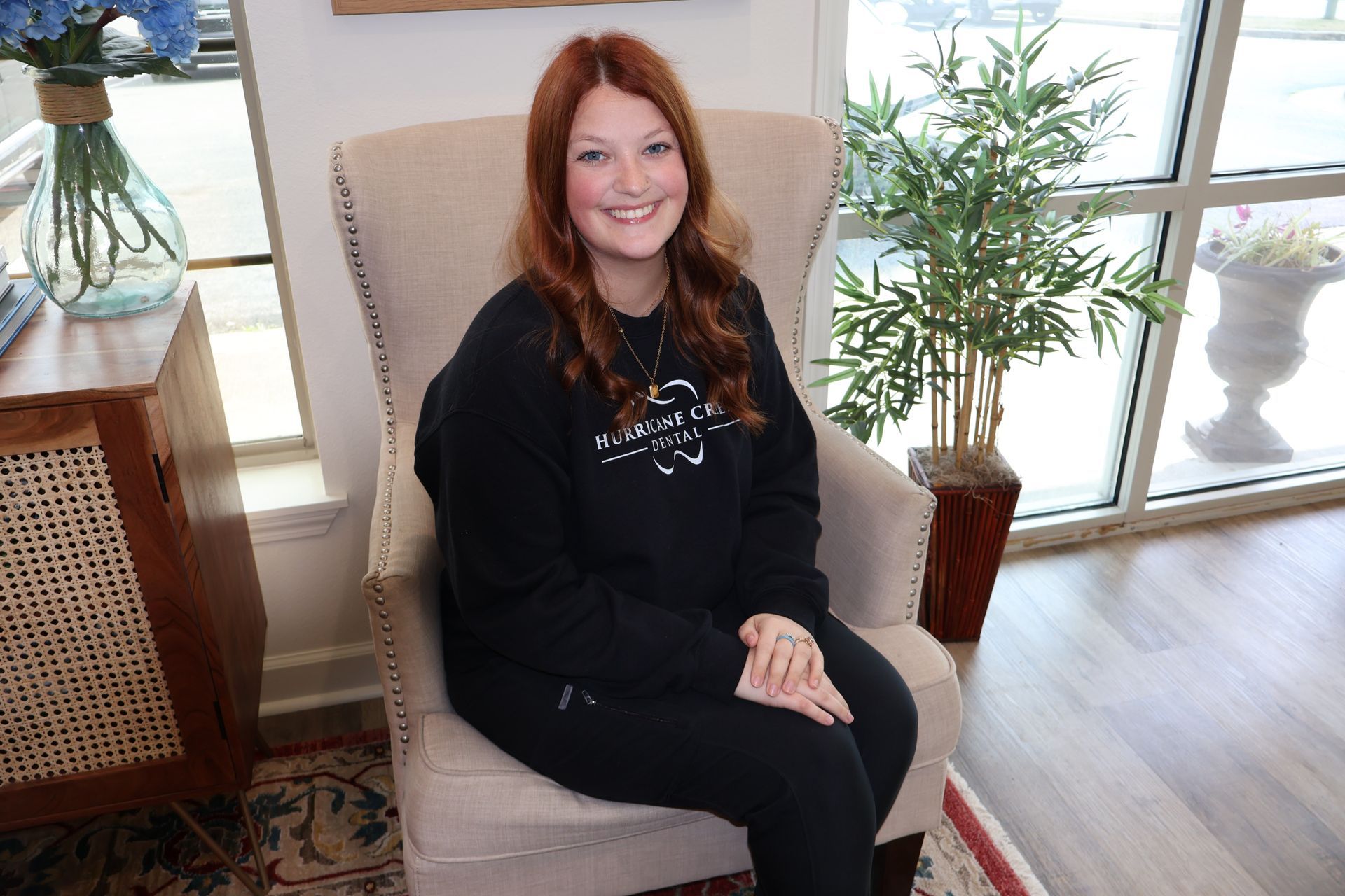 A smiling person with auburn hair sits in a beige wingback chair, wearing a black sweatshirt inside a sunlit office.