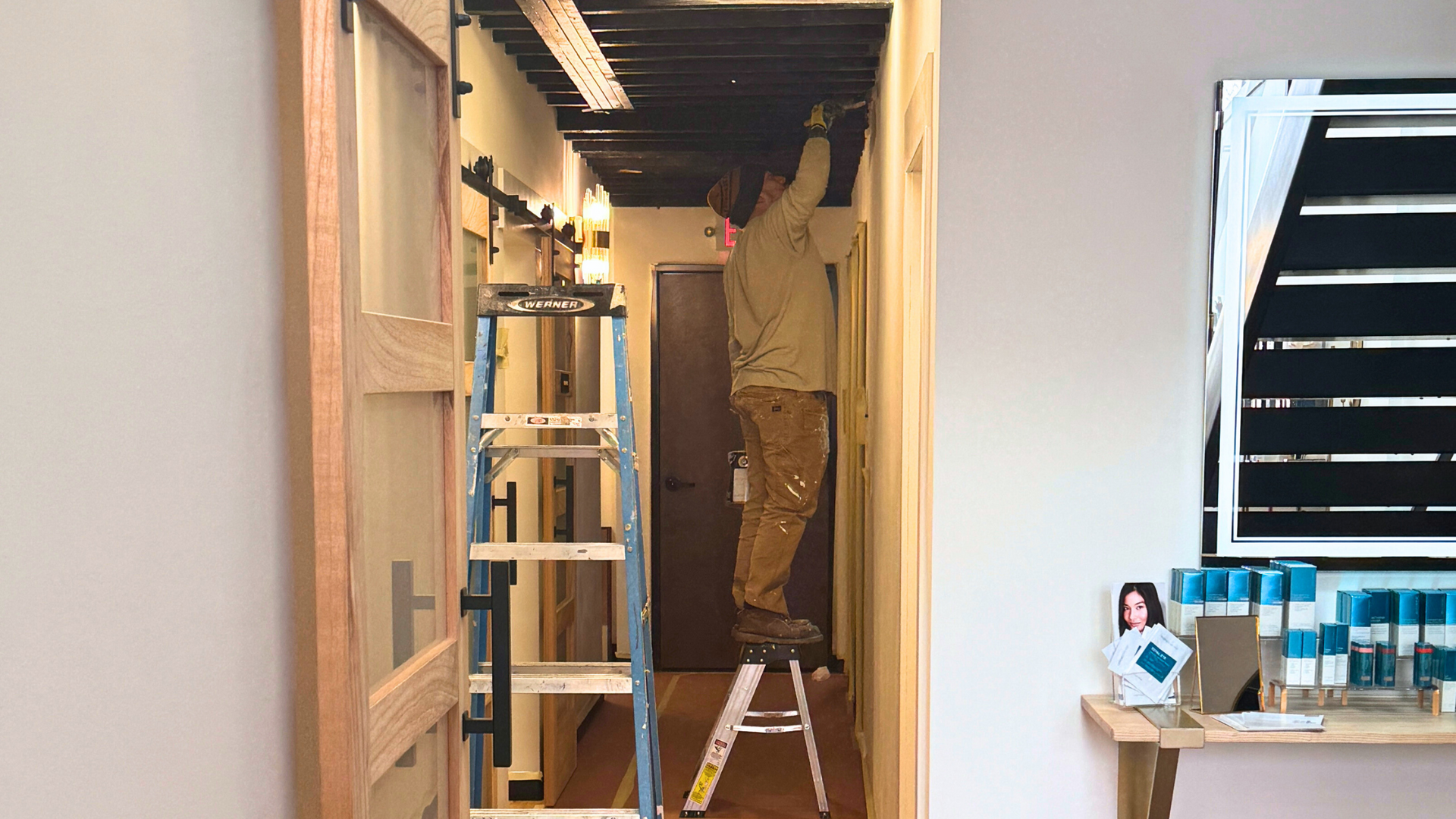 painter standing on a ladder, painting an open black ceiling in a modern hallway 



