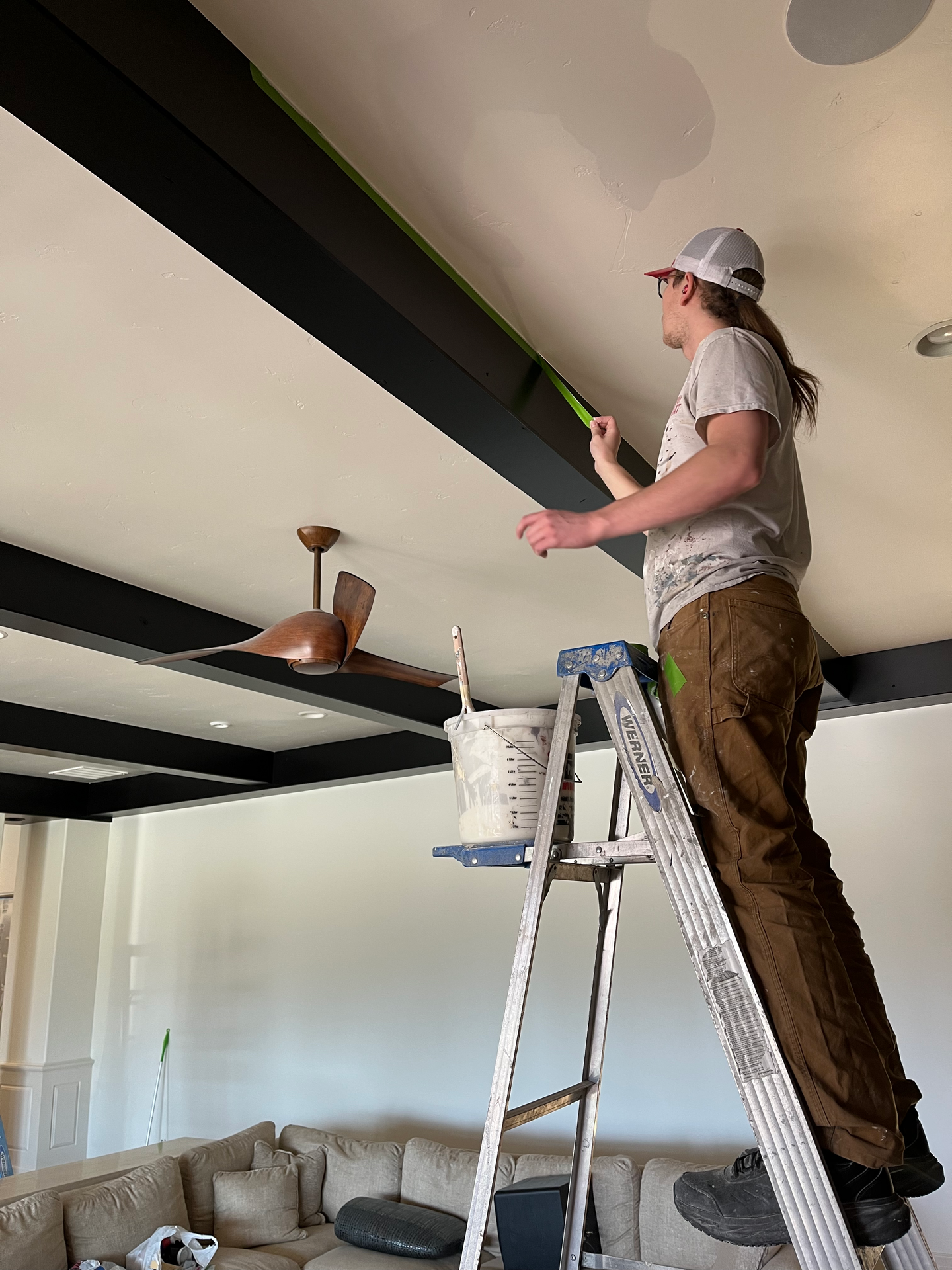 A man is standing on a ladder painting the ceiling of a living room.