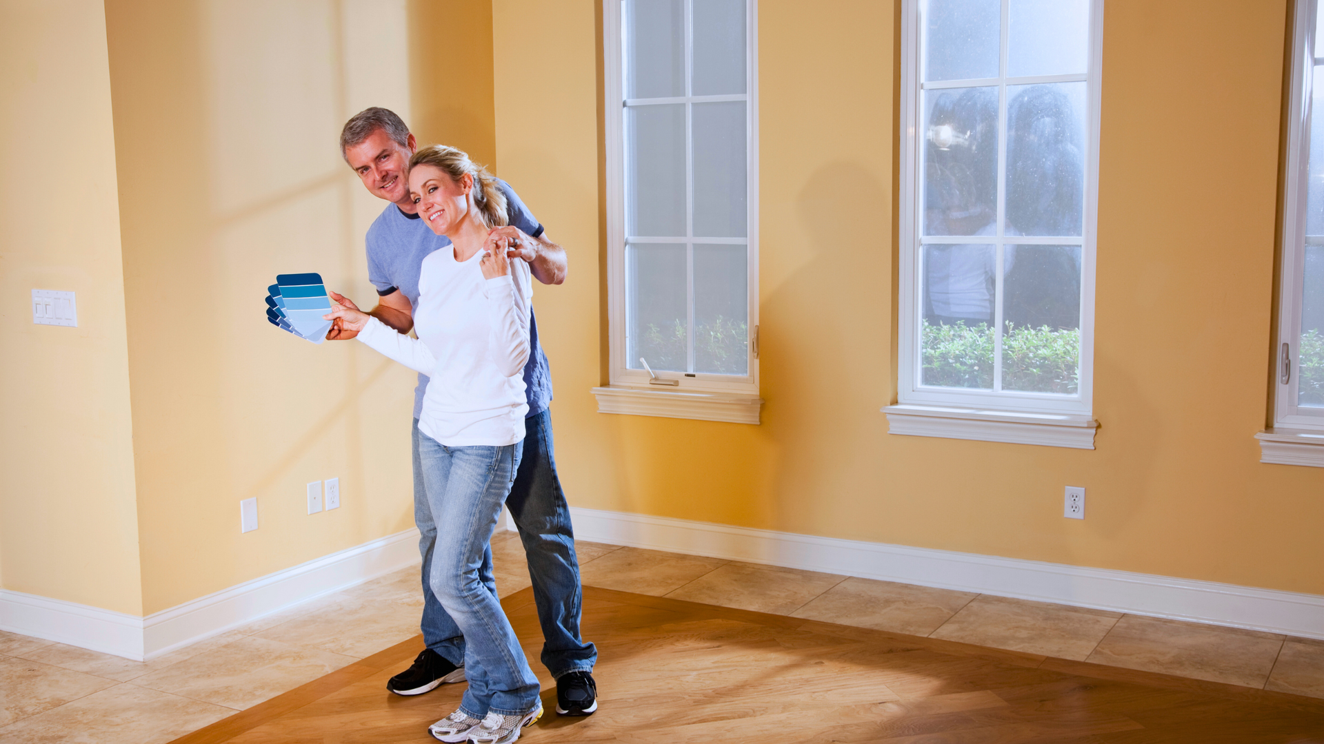 A man and woman are standing in an empty room looking at a paint swatch.