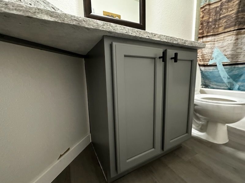 A bathroom with gray cabinets , a toilet , a sink and a mirror.