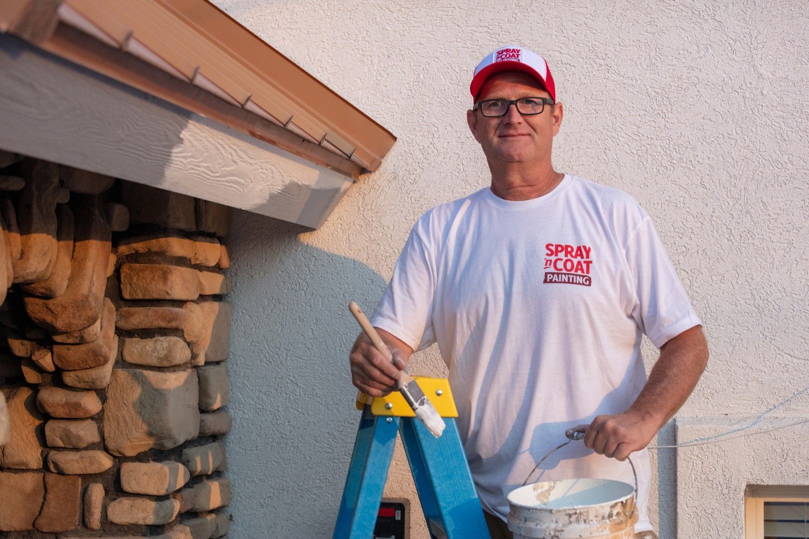 A man is standing on a ladder holding a bucket of paint and a brush.