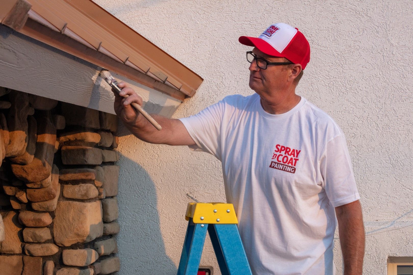 A man is standing on a ladder painting a stone wall.