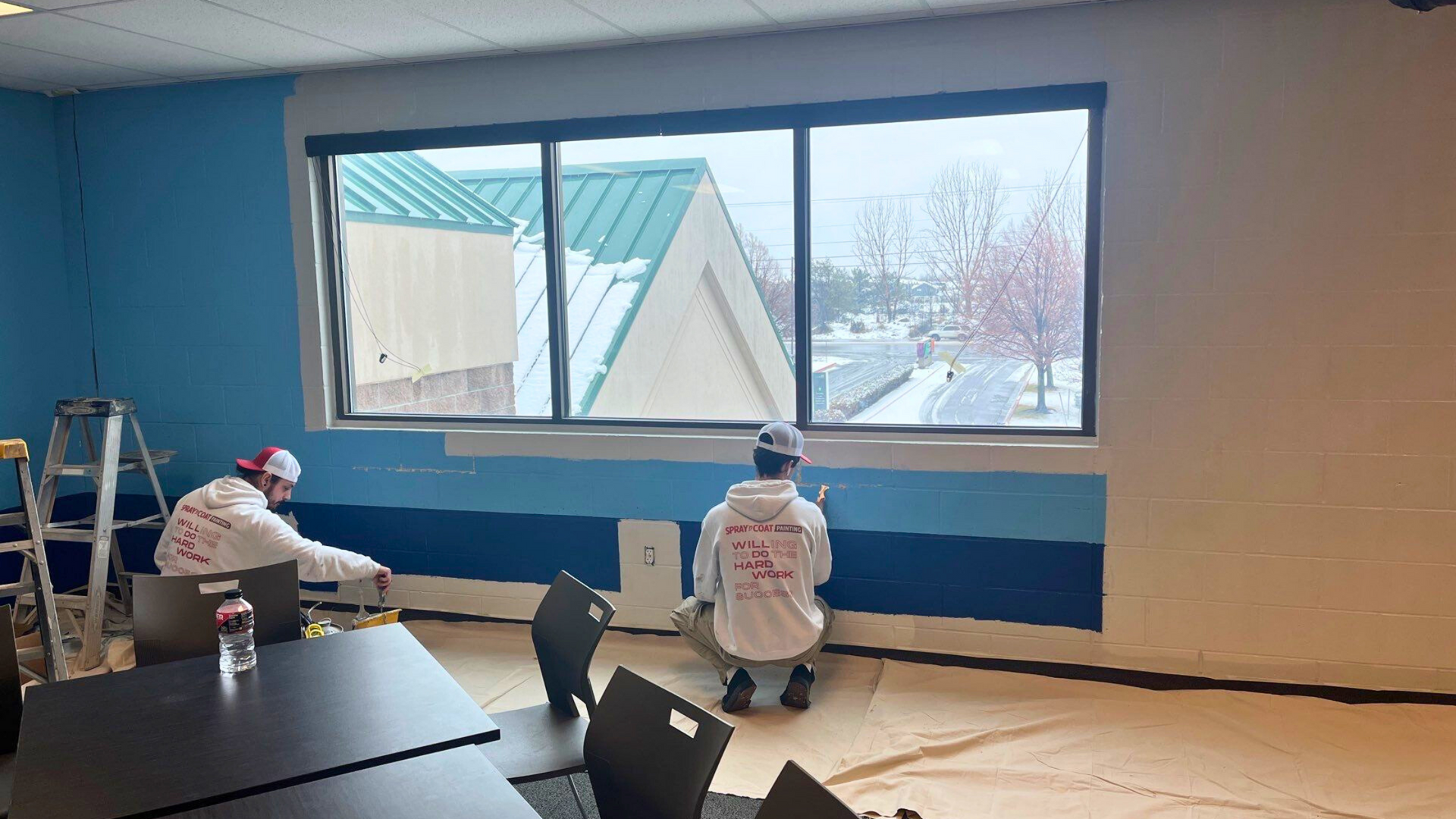 Two Spray 'n Coat painters apply blue paint to a commercial wall, using rollers and brushes.
