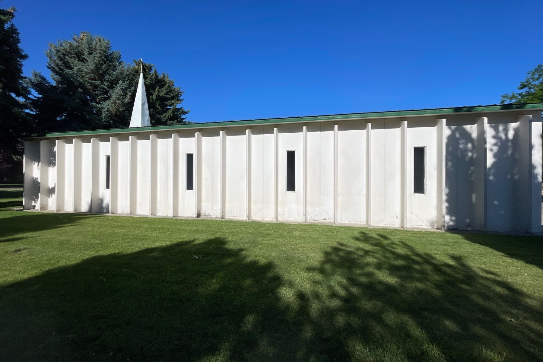 A white building with a green roof is surrounded by grass and trees