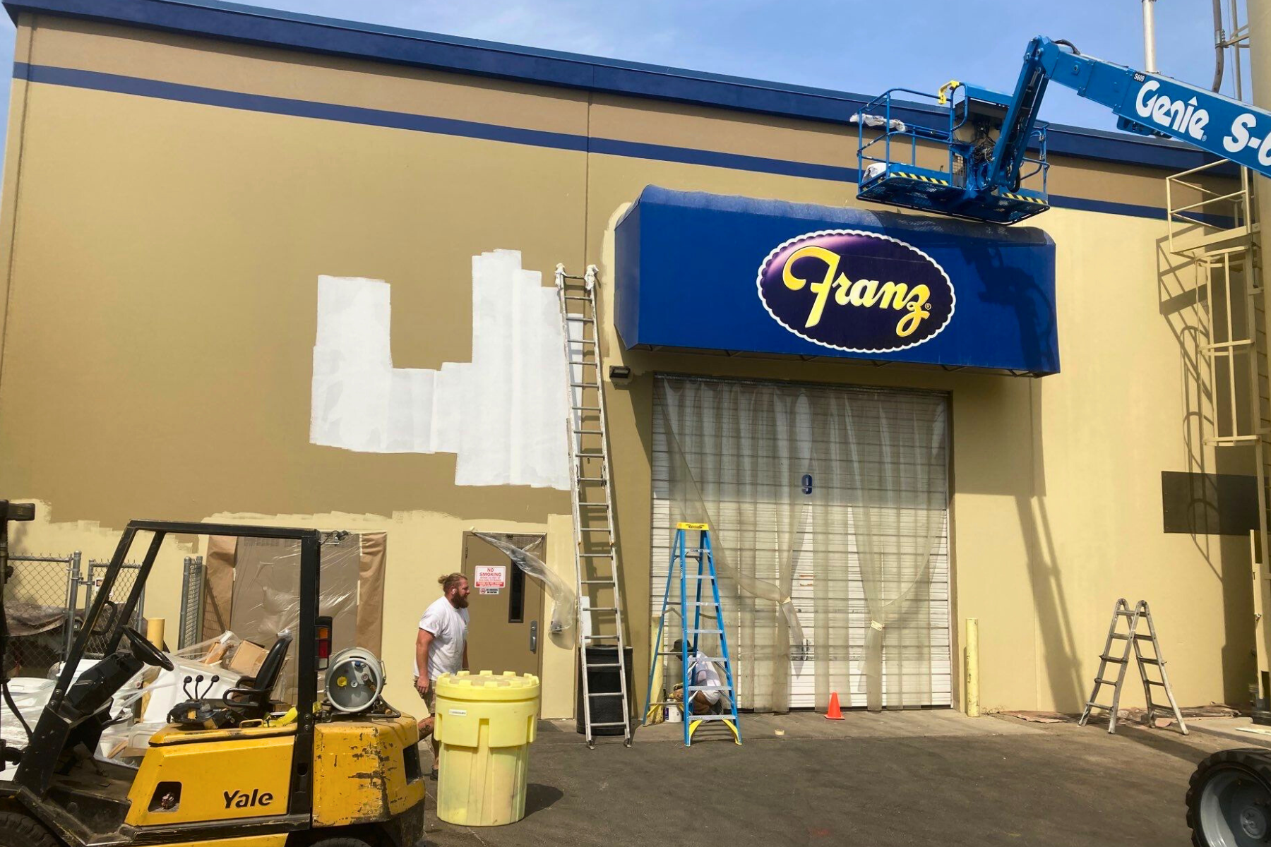 A yellow forklift is parked in front of a building that is being painted.