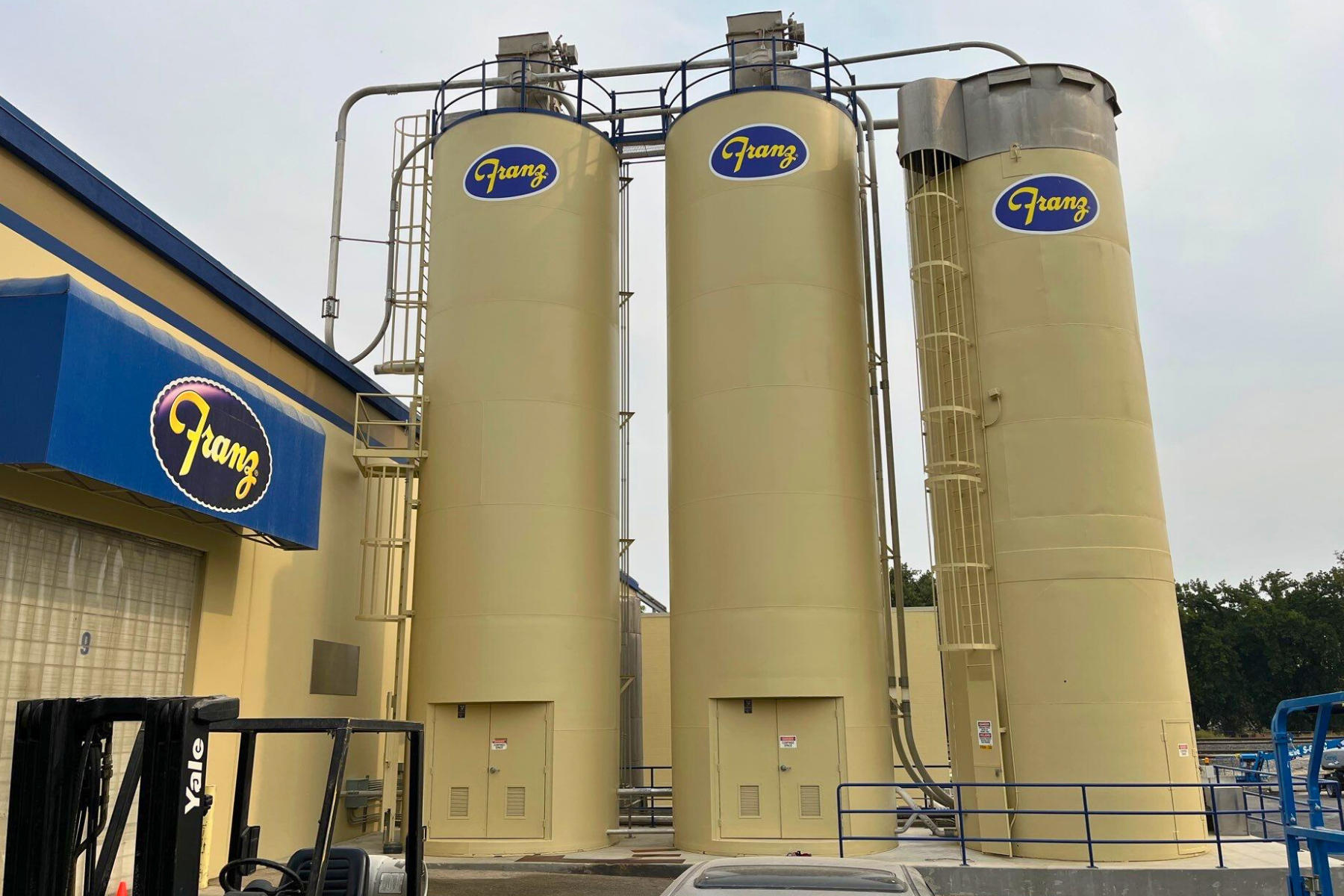 A forklift is parked in front of a building with three silos.