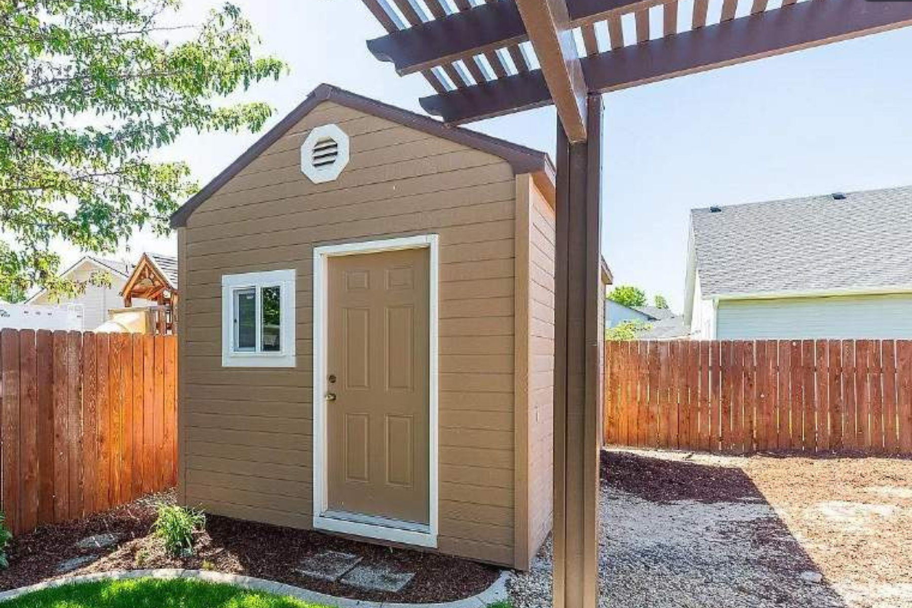 A small brown shed is sitting under a pergola in a backyard.