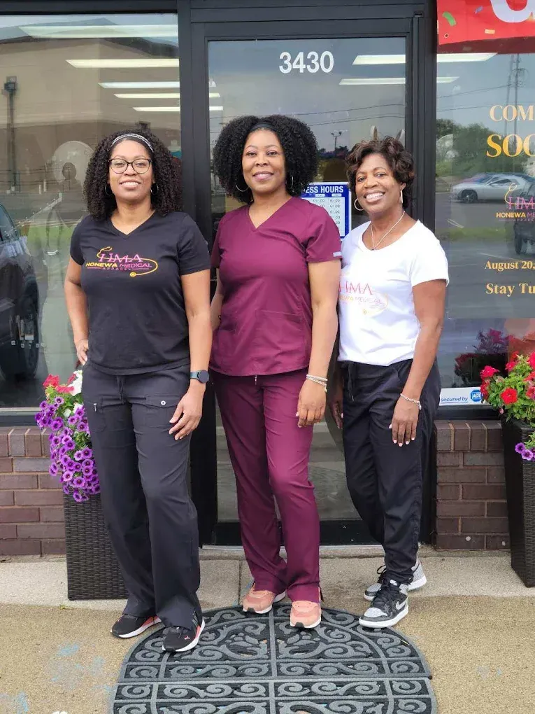 Three women stand outside a business entrance. One wears a black shirt and pants, another a maroon uniform, and the third a white shirt and black pants.