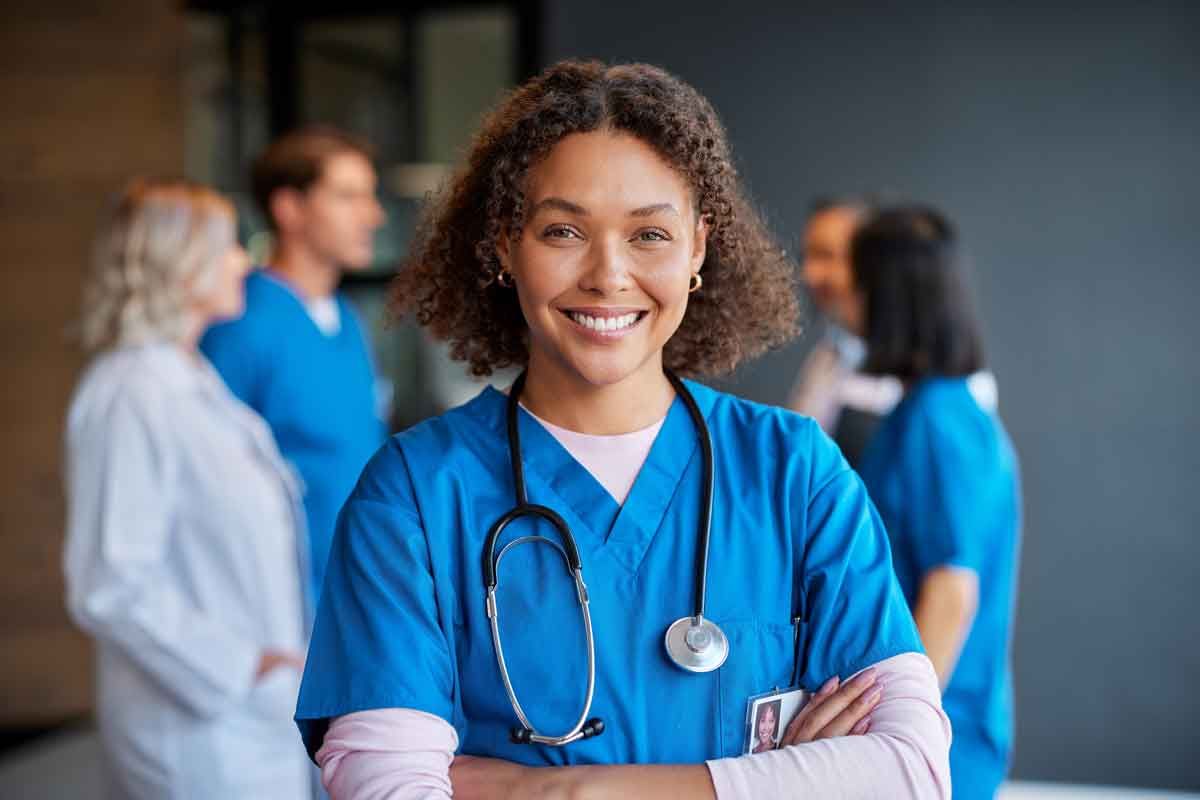 Smiling healthcare worker in blue scrubs with stethoscope, arms crossed; colleagues blurred in background.
