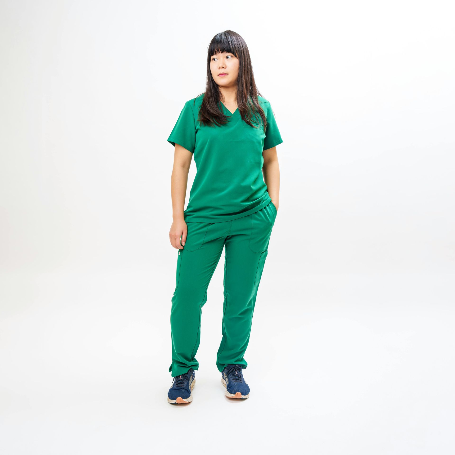 Woman in green scrubs stands with hands in pockets against a white backdrop.