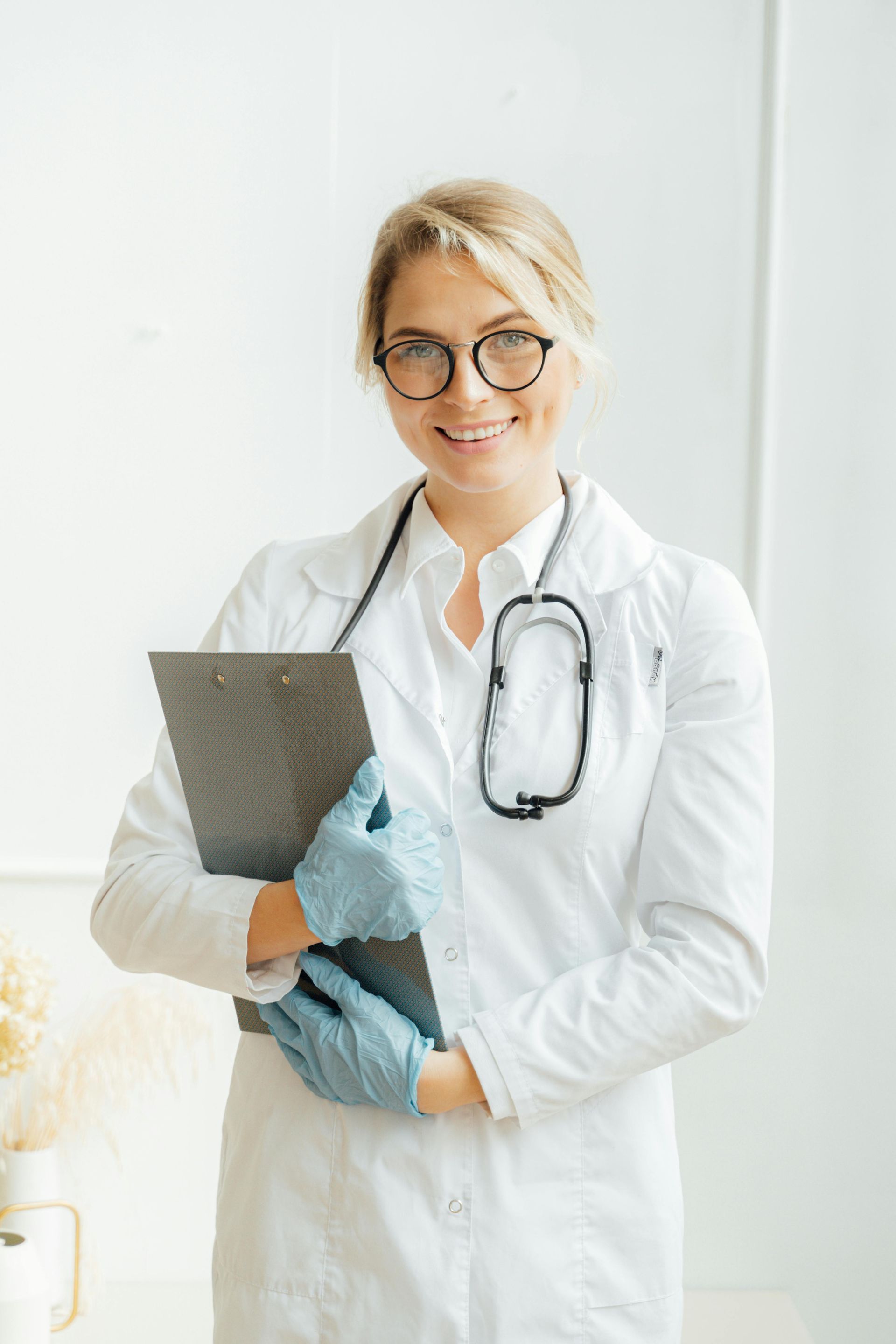 Doctor in white coat, stethoscope, and glasses, holding clipboard, smiling.