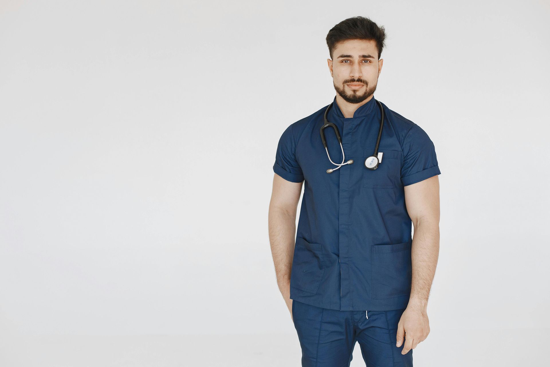 Man in blue scrubs with a stethoscope around his neck, hands in pockets, standing against white background.