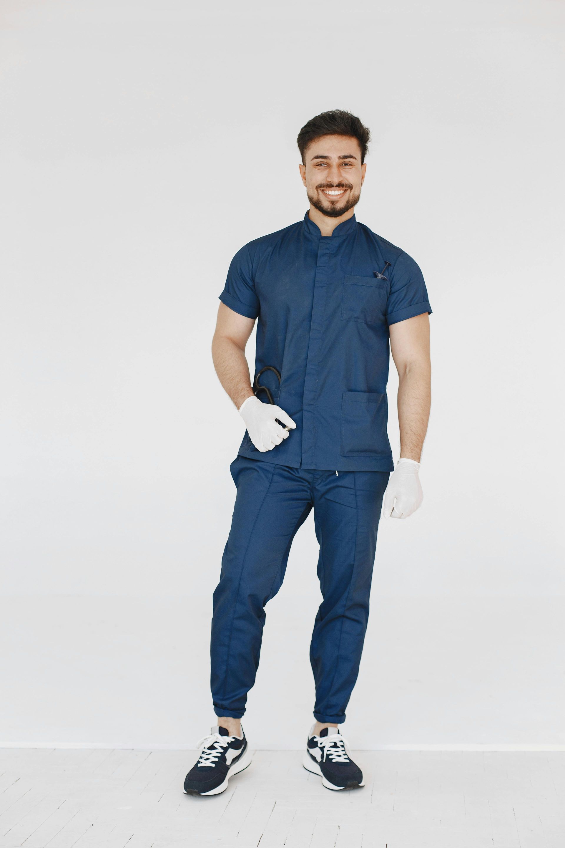 Man in blue scrubs, gloves, and sneakers smiles against a white background.