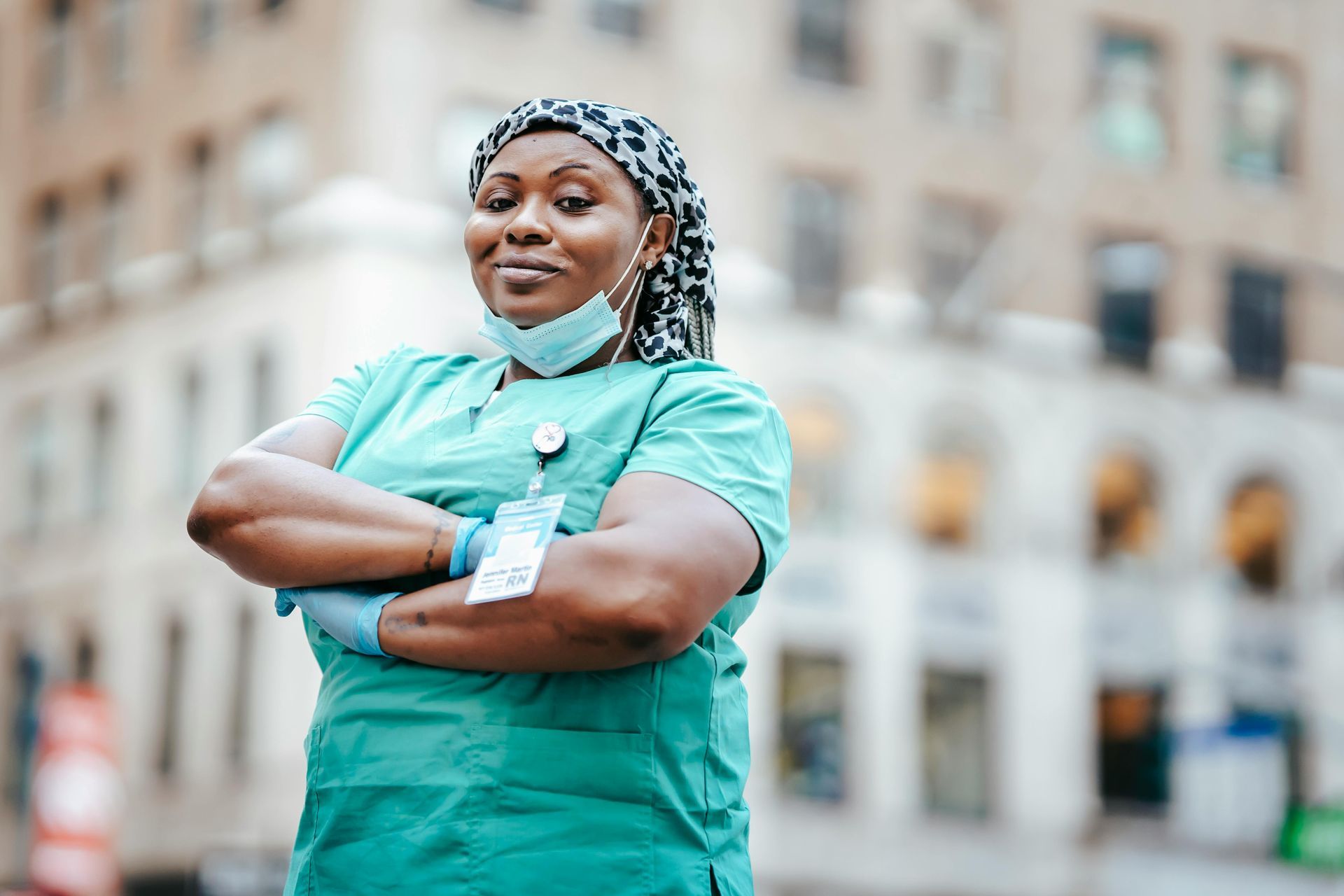 Medical worker in turquoise scrubs, arms crossed, standing outdoors in front of a building.
