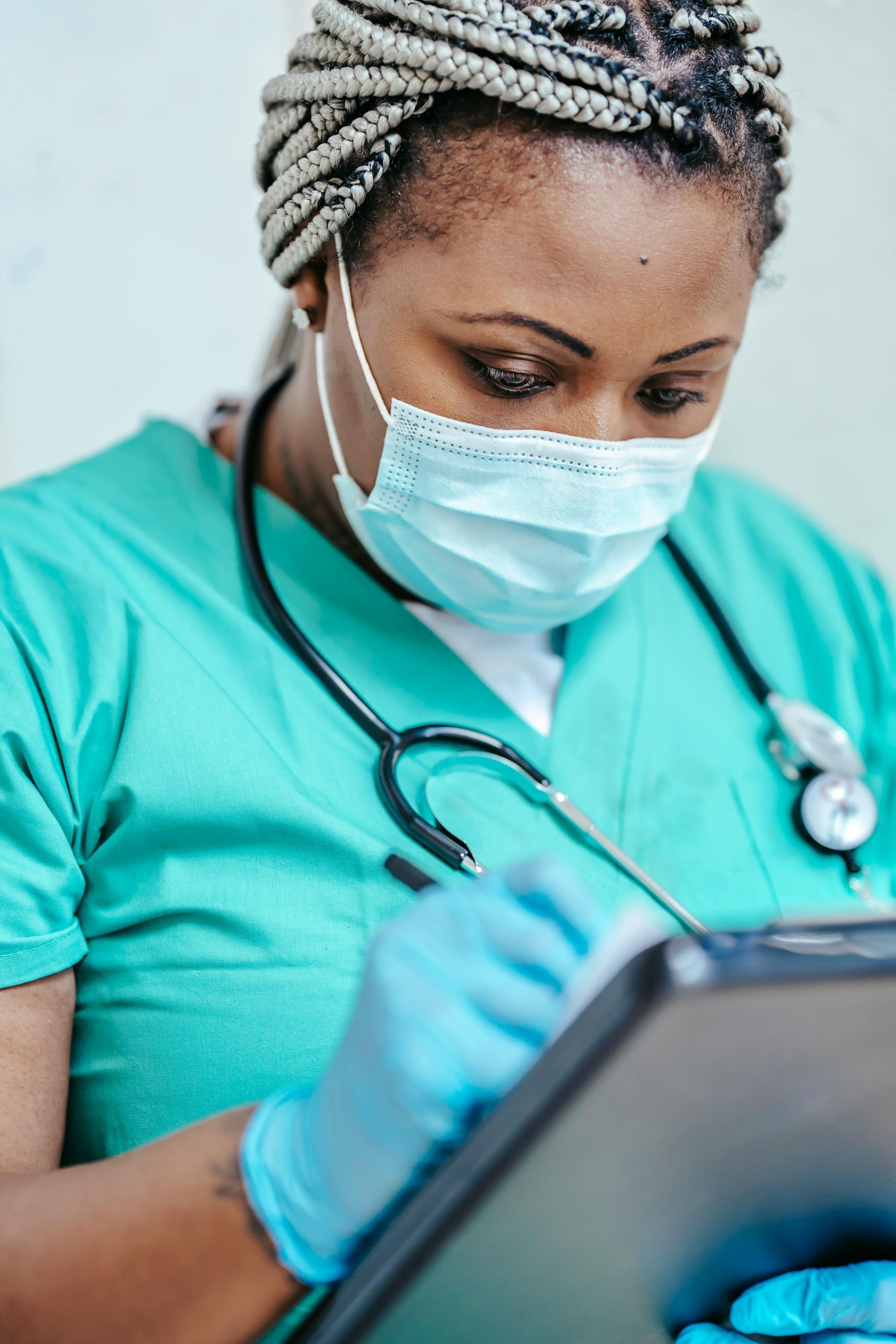 Nurse in teal scrubs, mask, and gloves, writing on a clipboard outdoors.