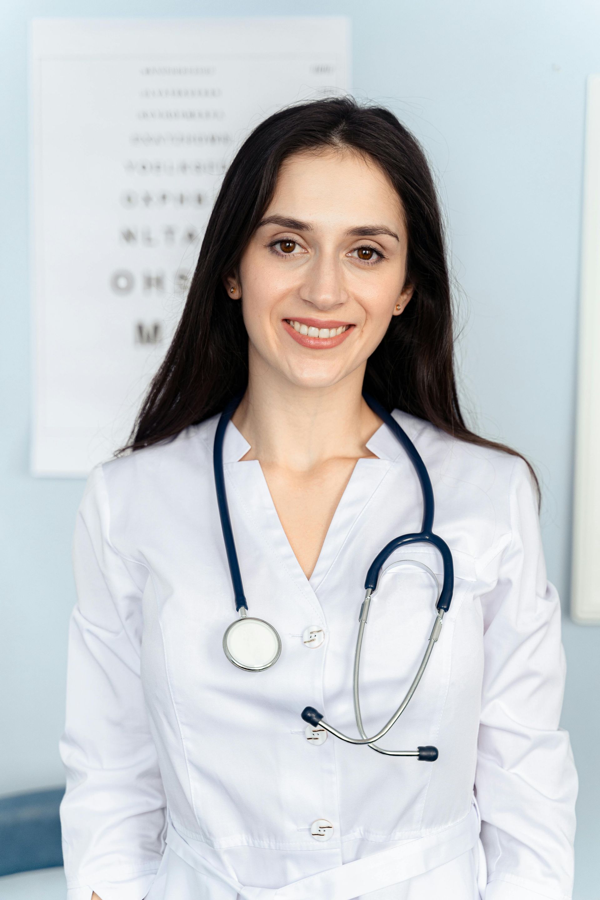 Woman in a white coat with a stethoscope smiling in an eye exam room.