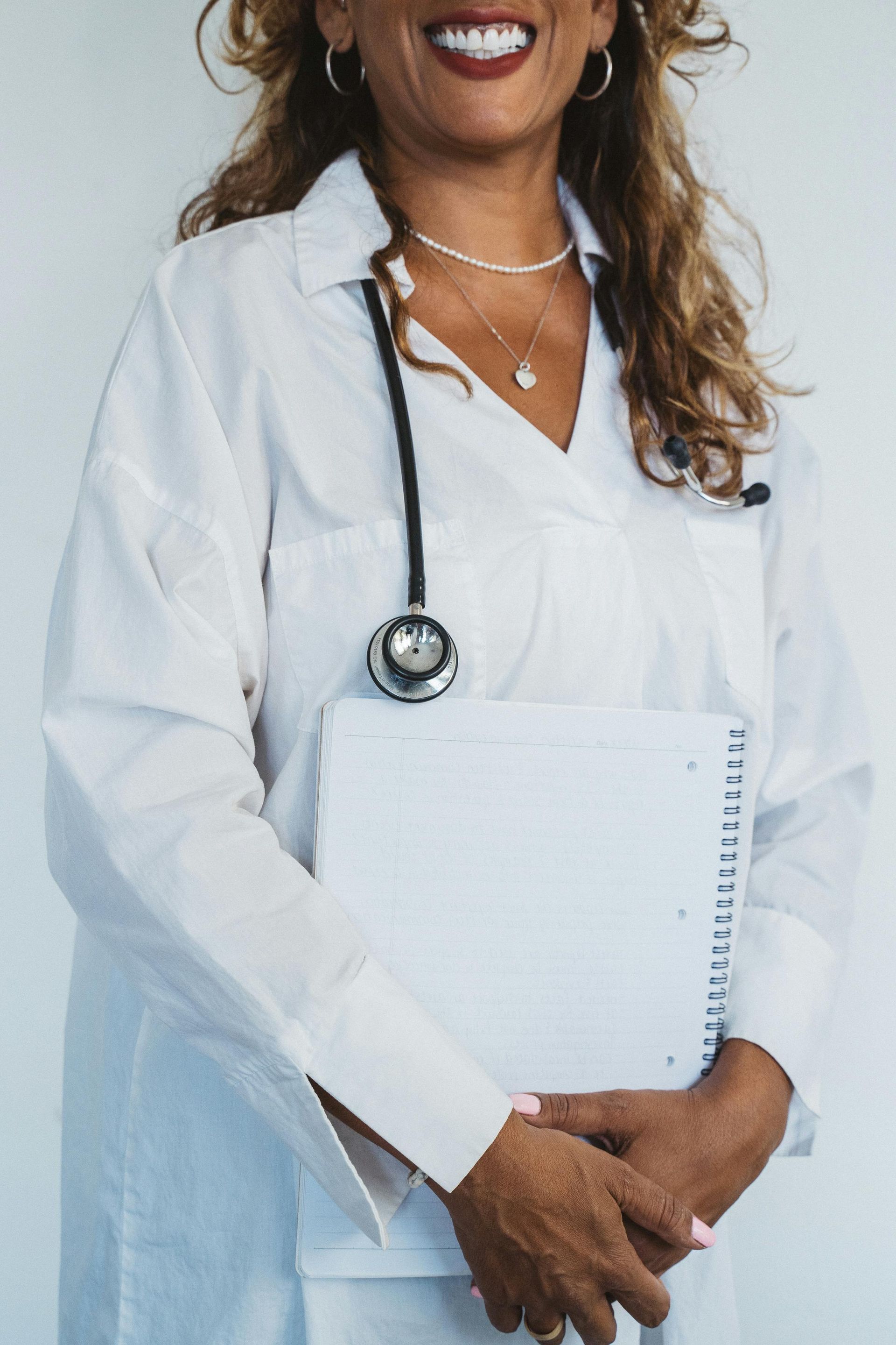Woman in white coat holding notepad and stethoscope, smiling.