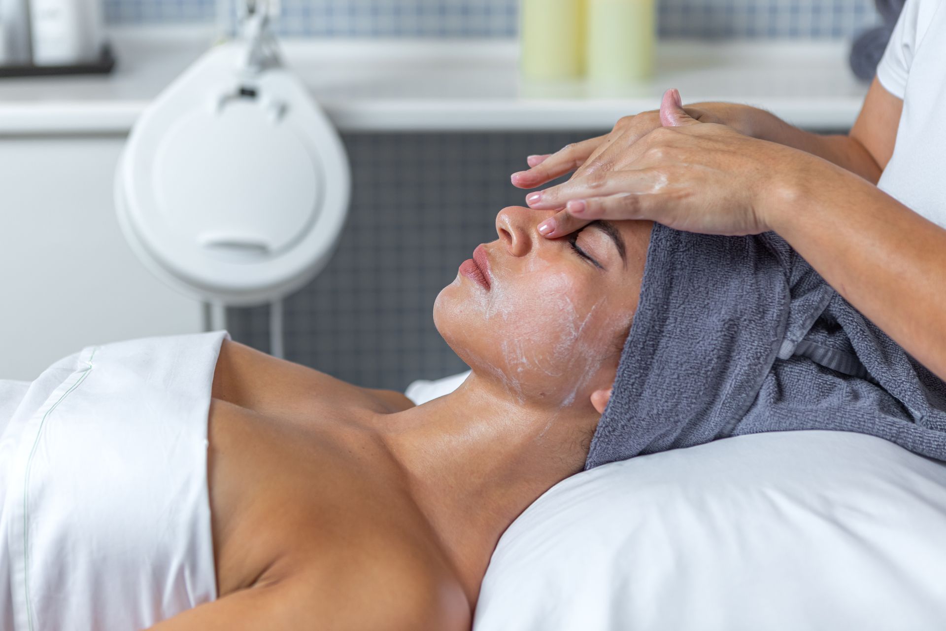A woman is getting a facial treatment at a spa with a towel wrapped around her head.