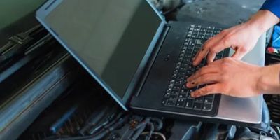 a man is typing on a laptop computer while working on a car engine .
