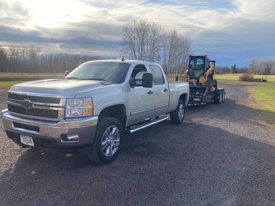 A silver truck is towing a yellow forklift down a dirt road.