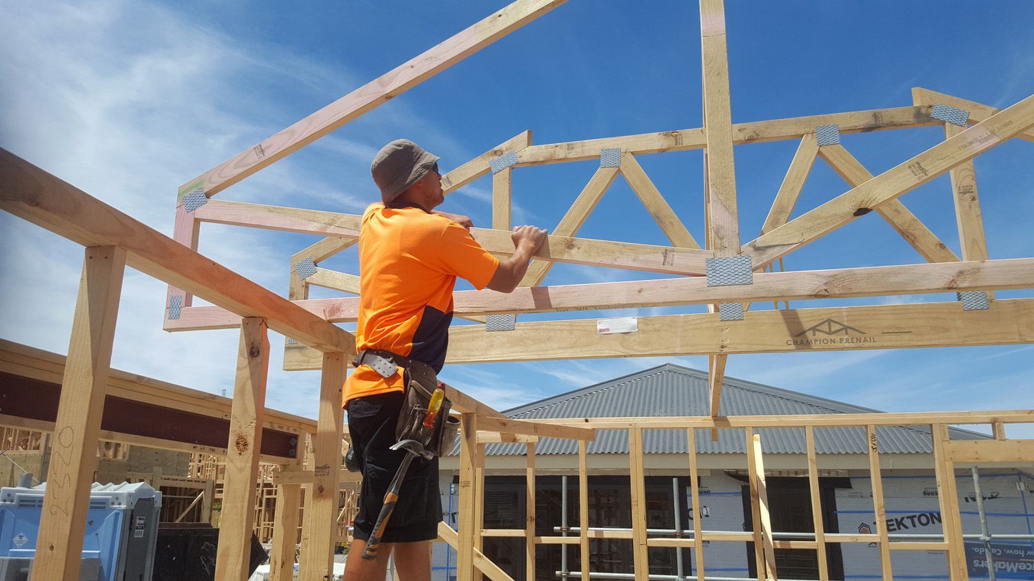 Construction worker building roof truss on a sunny day. Wearing orange shirt, standing on scaffolding.
