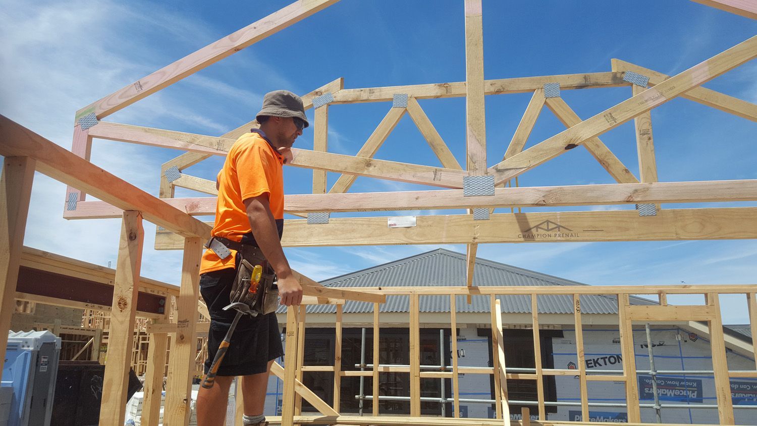 Carpenter on a wooden structure, installing beams. Bright orange shirt, sunny sky.
