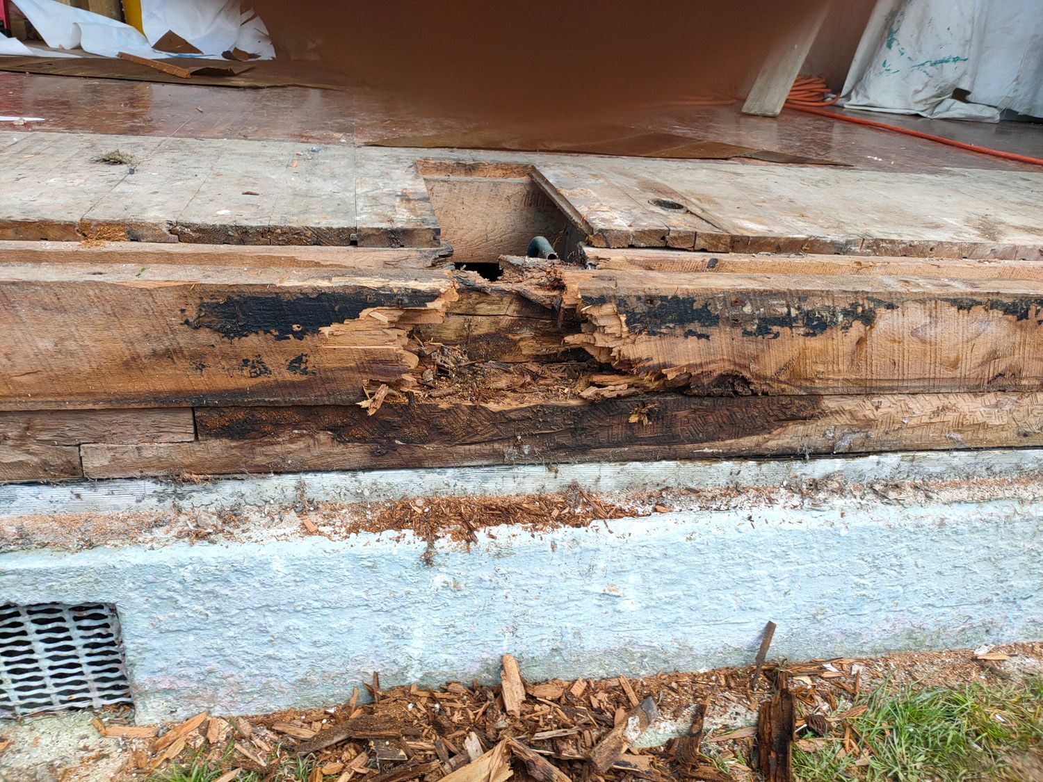 Damaged wooden foundation of a building with visible rot and decay, near a concrete base.
