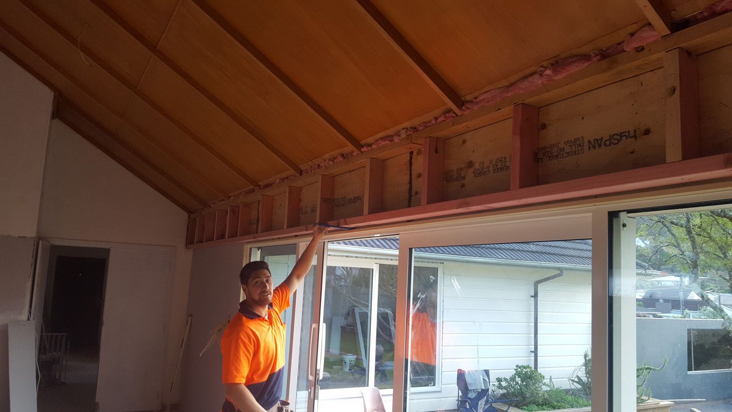 Man in an orange shirt points to wooden framing above large windows, interior construction site.