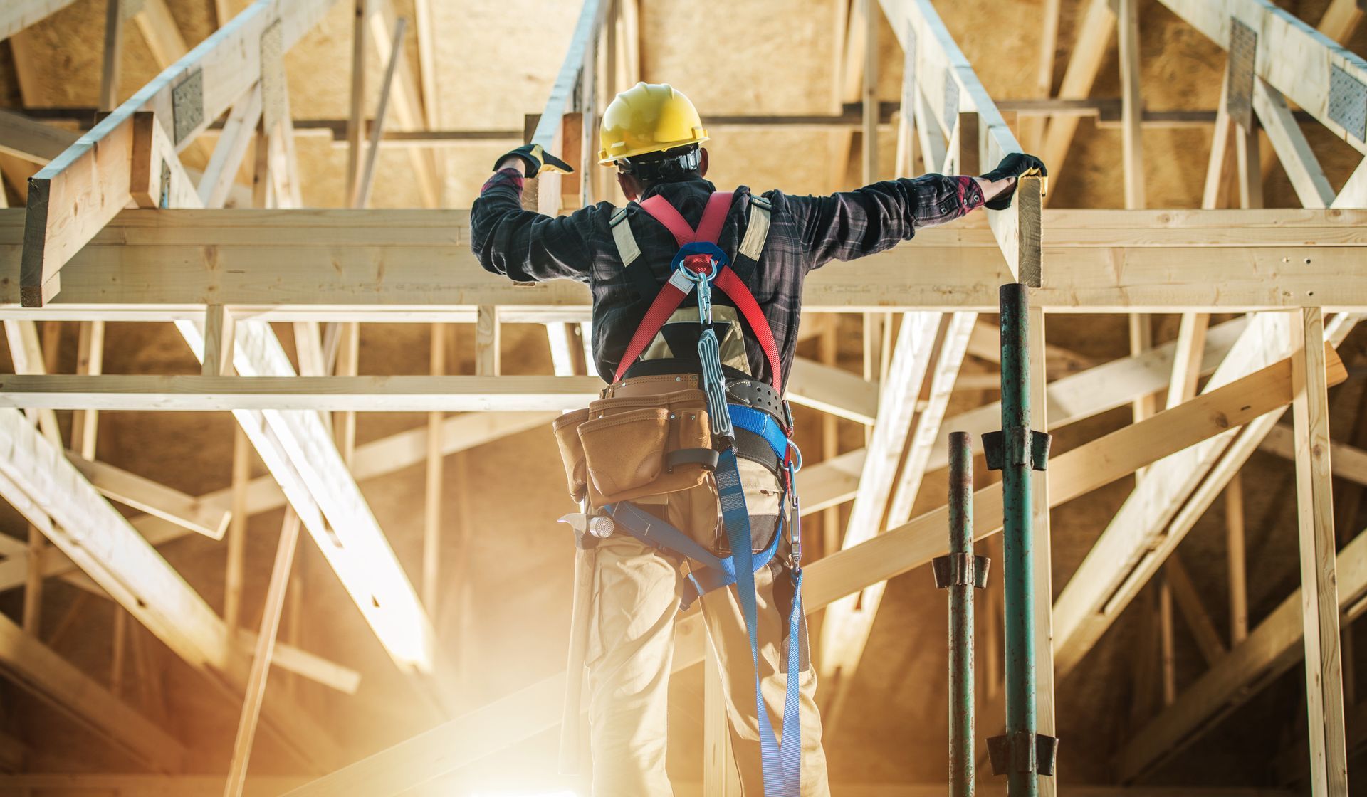 Construction worker on roof frame wearing safety harness, hammering wood, sunny interior.