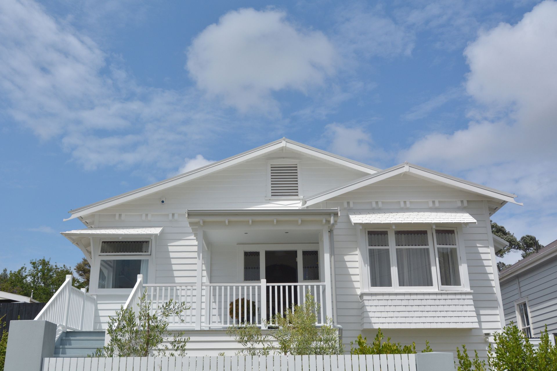 White two-story house with a porch and shutters under a blue sky with clouds.