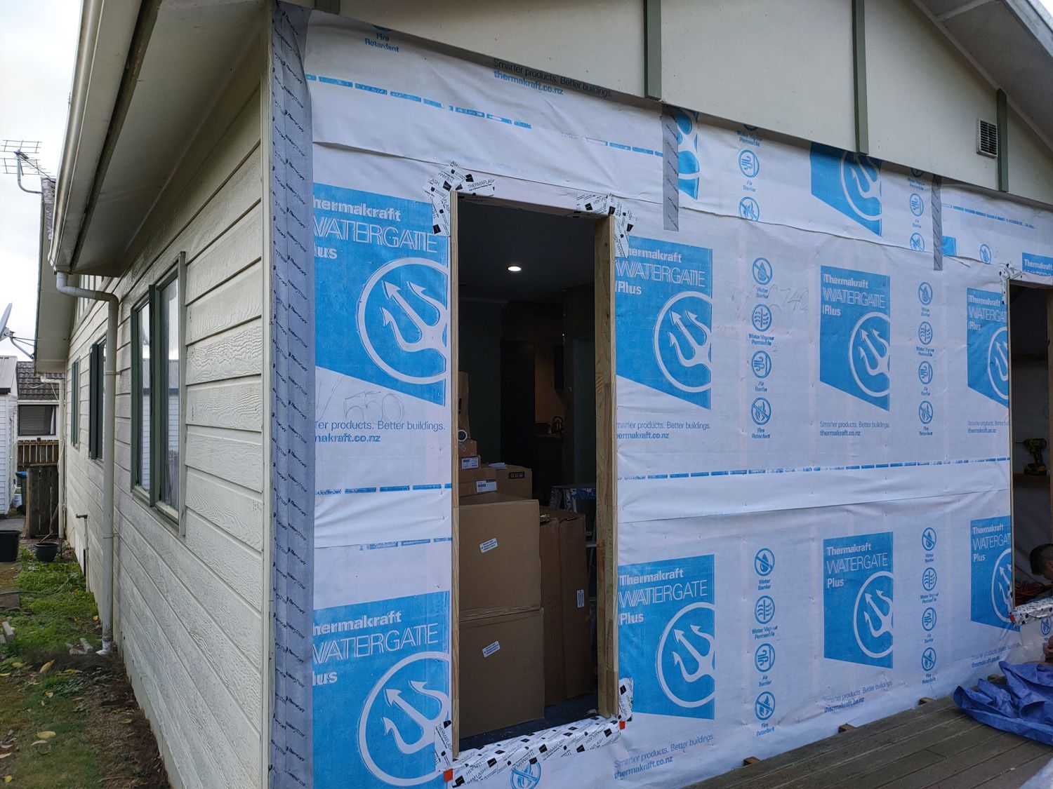 Exterior wall of a building under construction, showing new door frame, blue wrap, and existing siding.