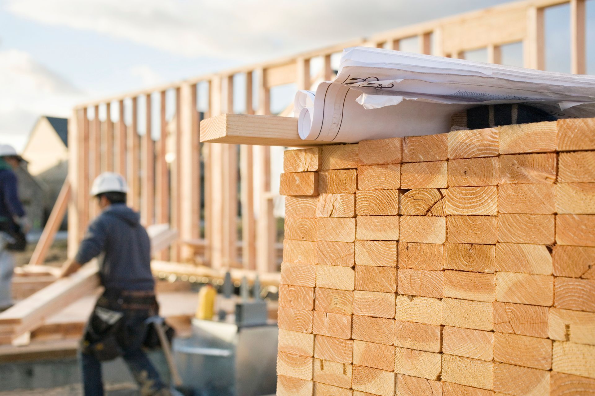 Construction workers building a wooden house; blueprints on lumber stack.