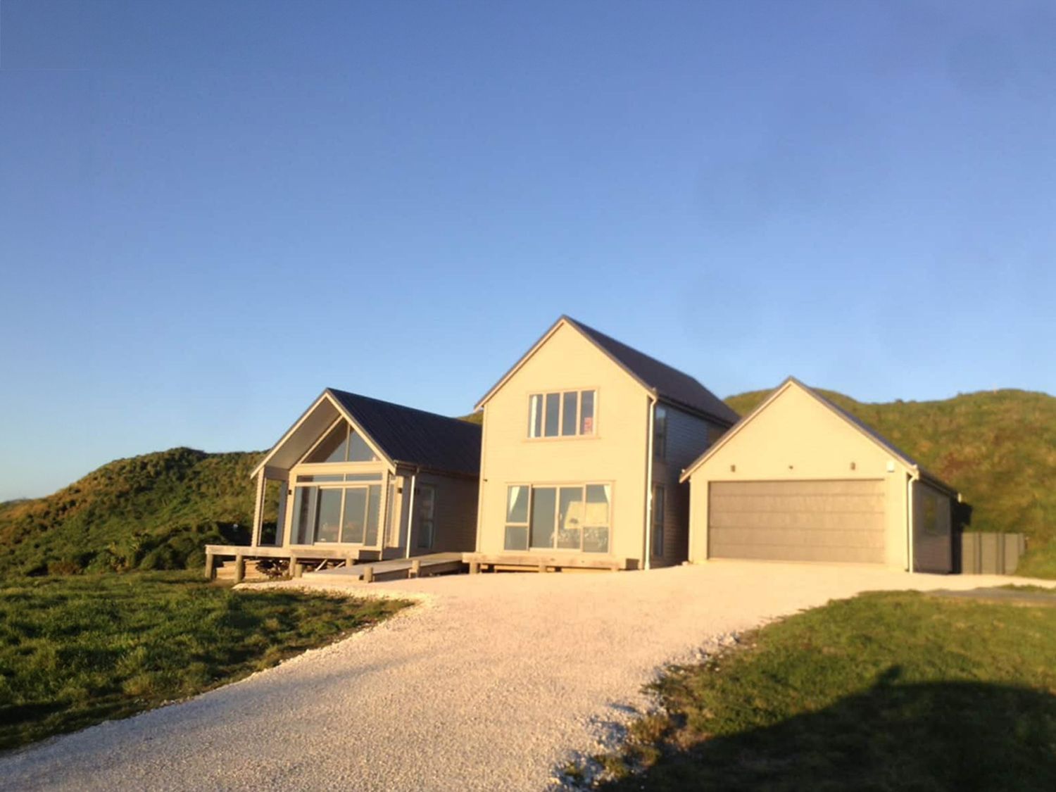 Modern house with light grey exterior, driveway, and garage against a grassy hill and blue sky.