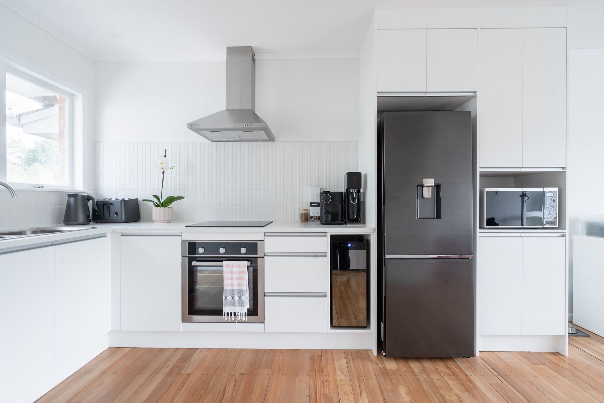 Modern white kitchen with stainless steel appliances, dark refrigerator, and wooden floors.