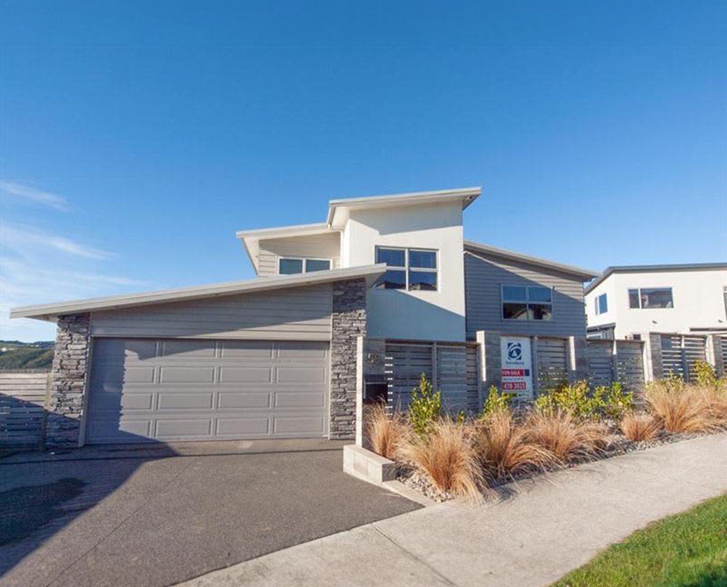 Modern two-story house with gray garage door, stone accents, and manicured landscaping under a blue sky.