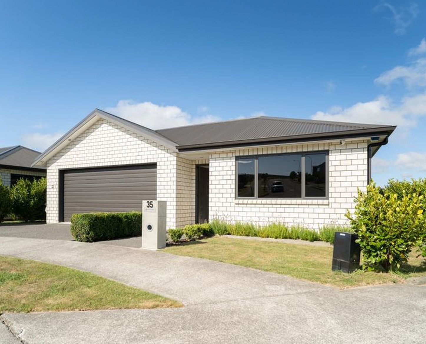 White brick house with a brown garage door, driveway, and shrubs under a blue sky.