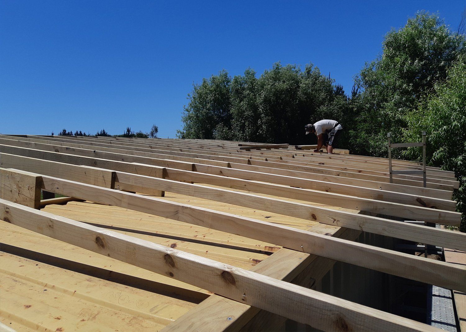 A person works on wooden beams. Bright blue sky, trees in the background.