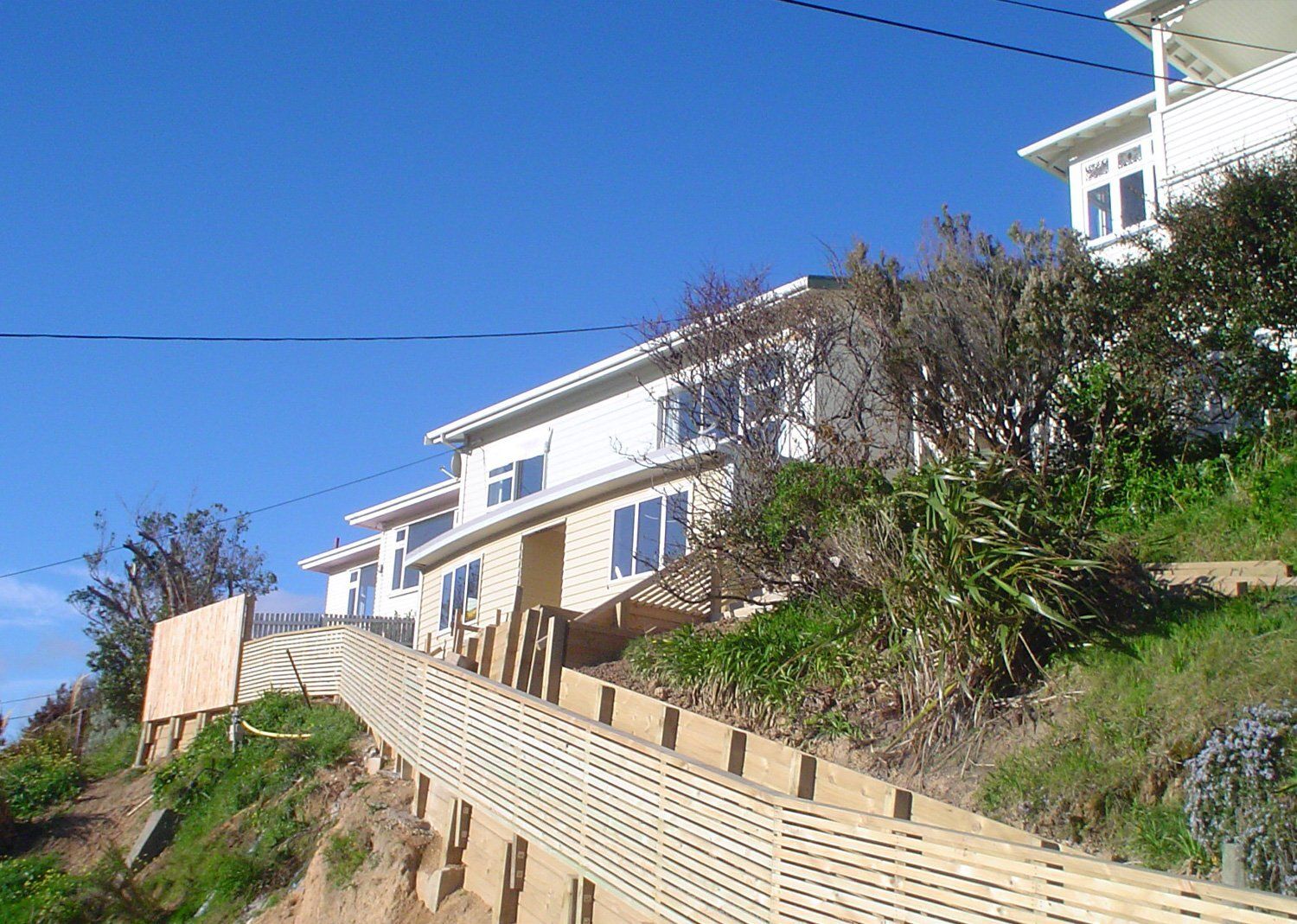 Two-story house on a steep hillside with wooden retaining walls and stairs under a bright blue sky.