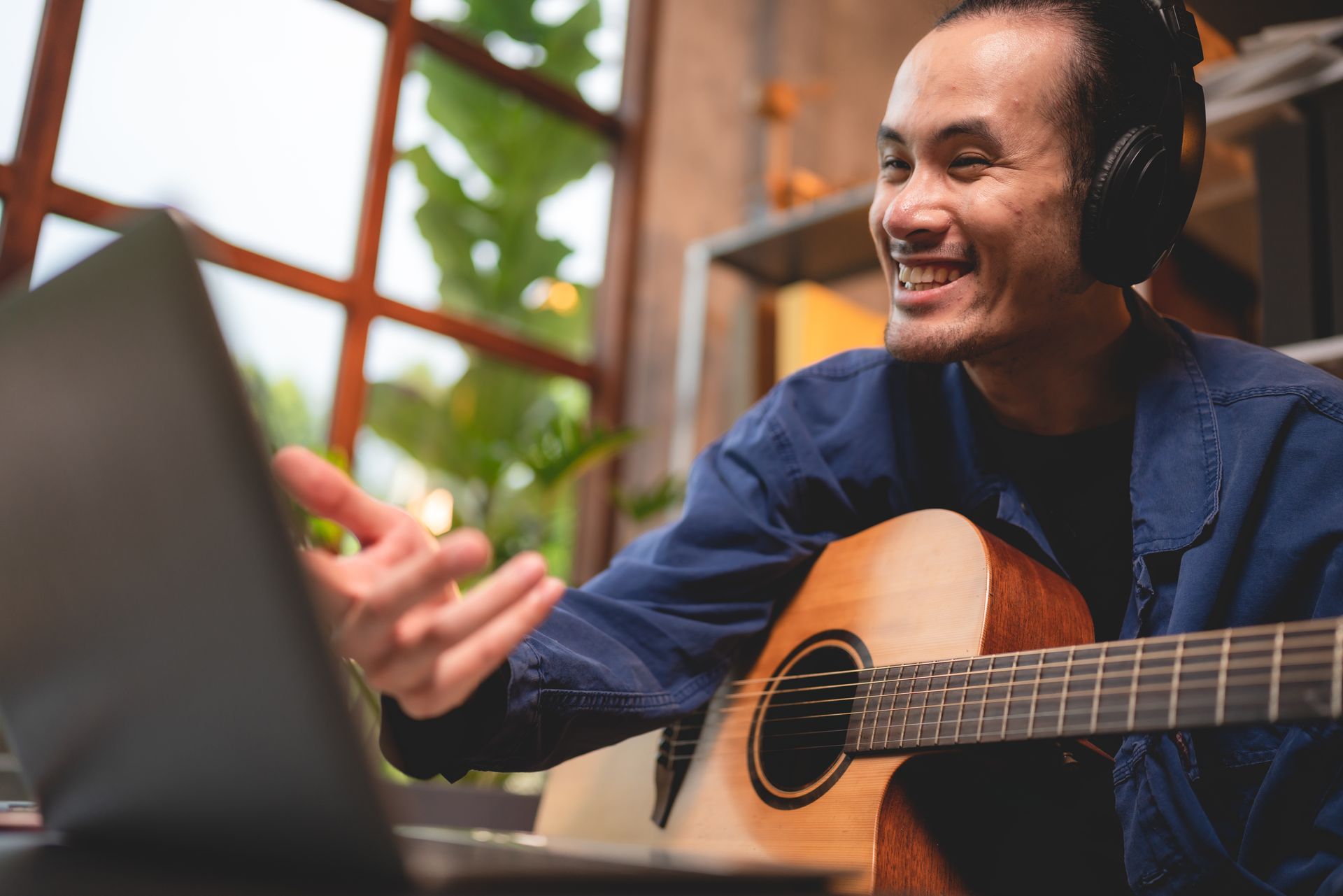 A man is playing an acoustic guitar in front of a laptop computer.