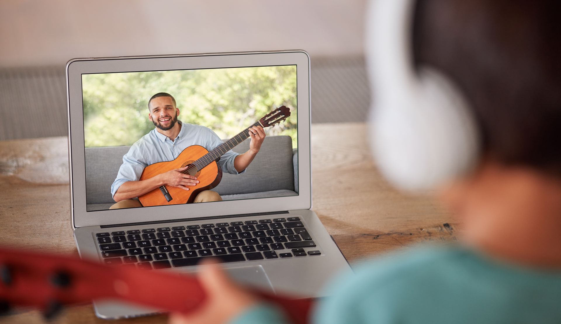 A child is playing a guitar while watching a video on a laptop.