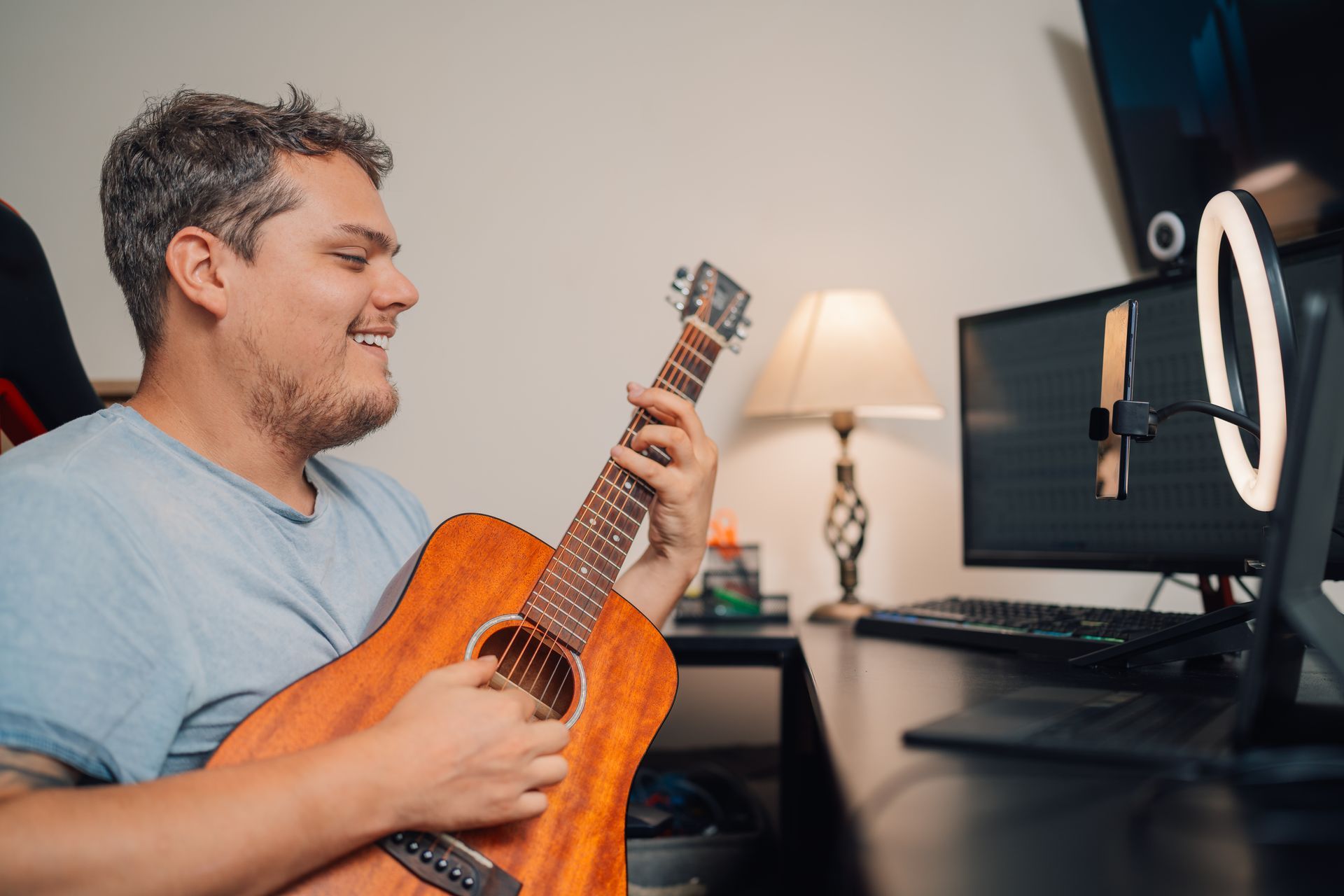 A man is playing an acoustic guitar in front of two computer monitors.