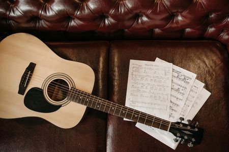 An acoustic guitar is sitting on a couch next to sheet music.