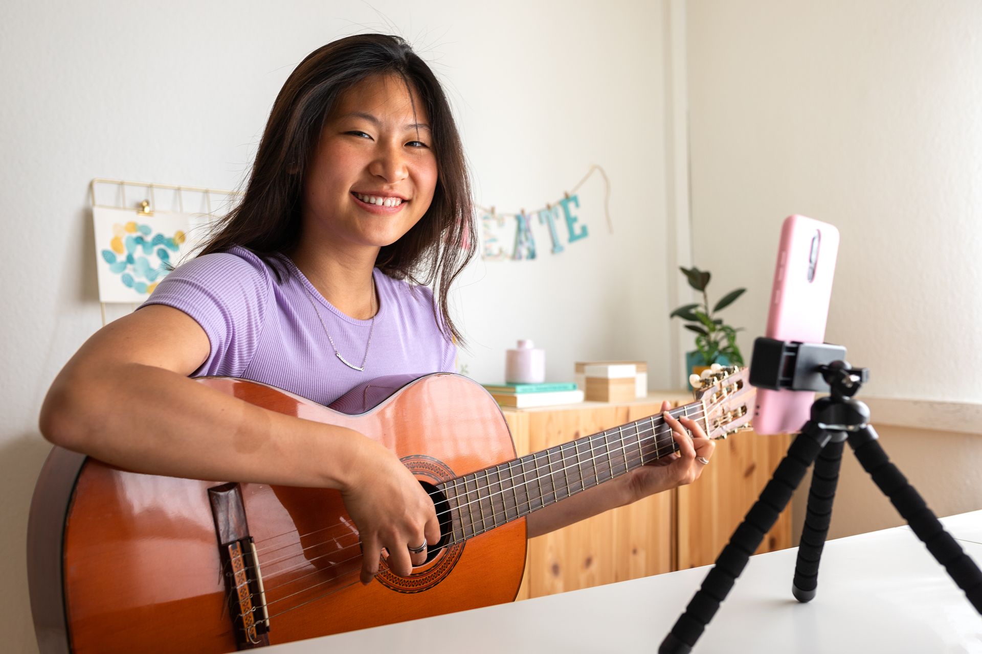 A woman is playing a guitar in front of a tripod and a cell phone.